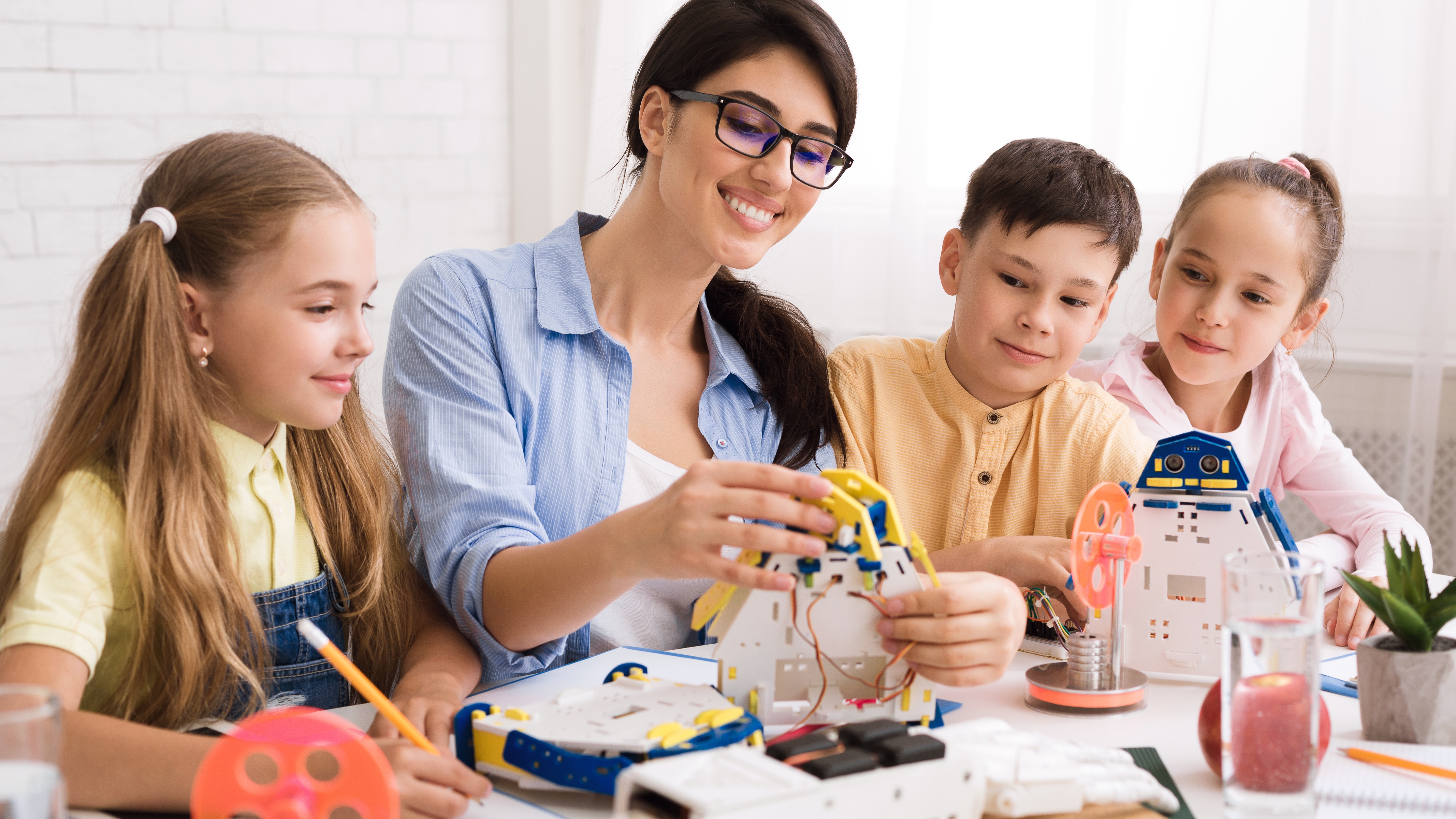 Woman teaching three children assembling robotic kits at a table in a bright room.