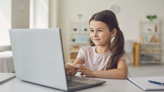 Young girl with pigtails sitting at a table and using a laptop in a bright room.