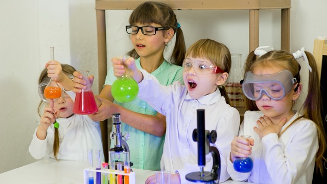 Four children wearing safety glasses holding colorful chemical liquids in a science lab.