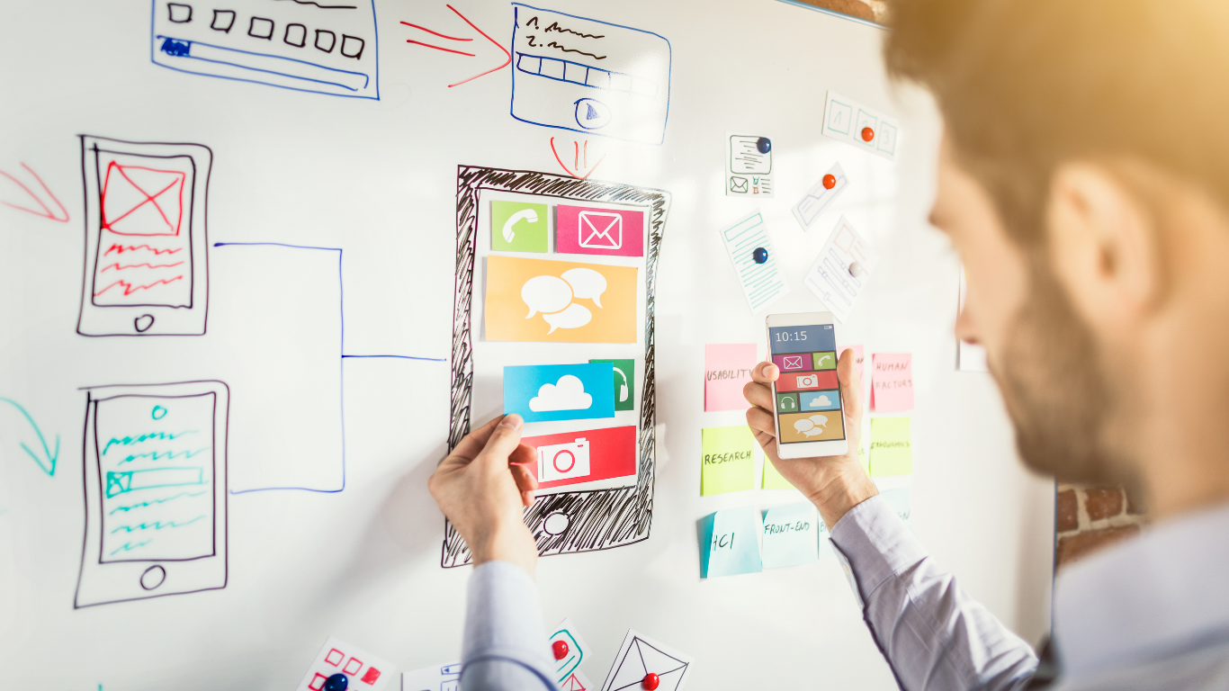 Person holding a smartphone and placing colored app icon cards on a whiteboard sketch of a mobile app interface.