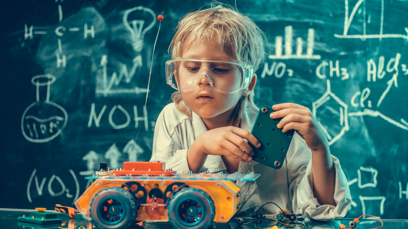 Child wearing safety goggles assembling a small robot with an orange chassis in front of a chalkboard with chemical formulas and diagrams.