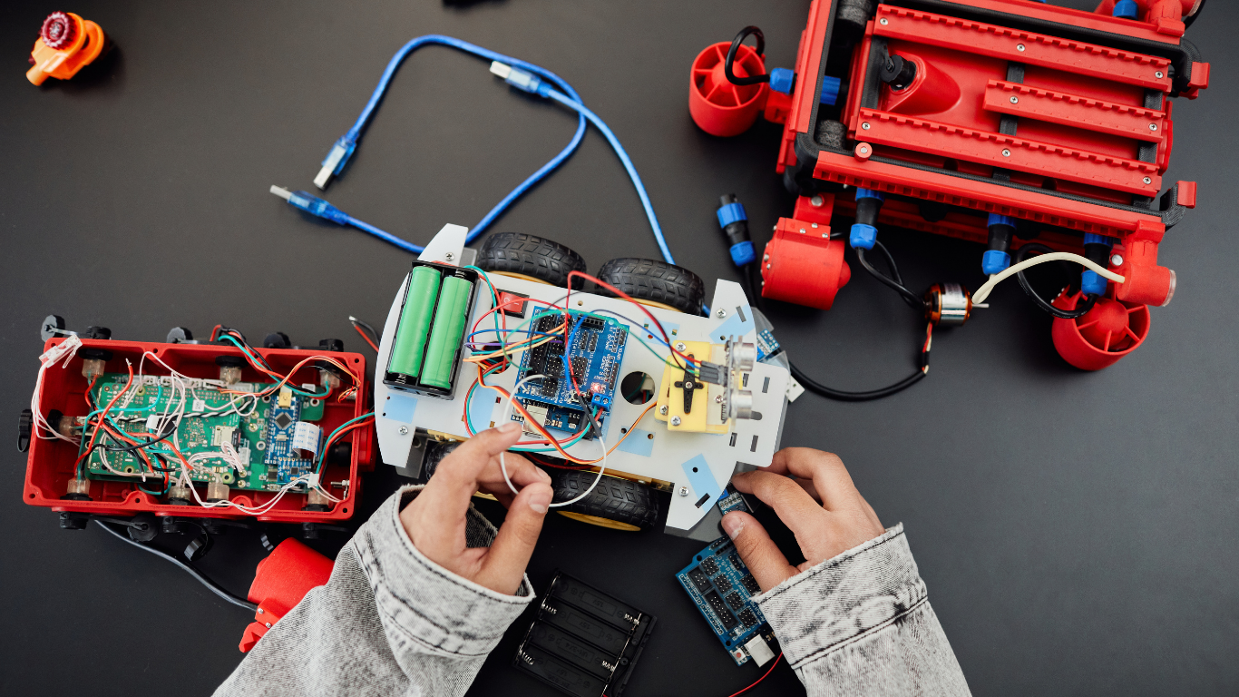 Person assembling electronic components and wiring on a small robotic vehicle with red and white parts on a black surface.