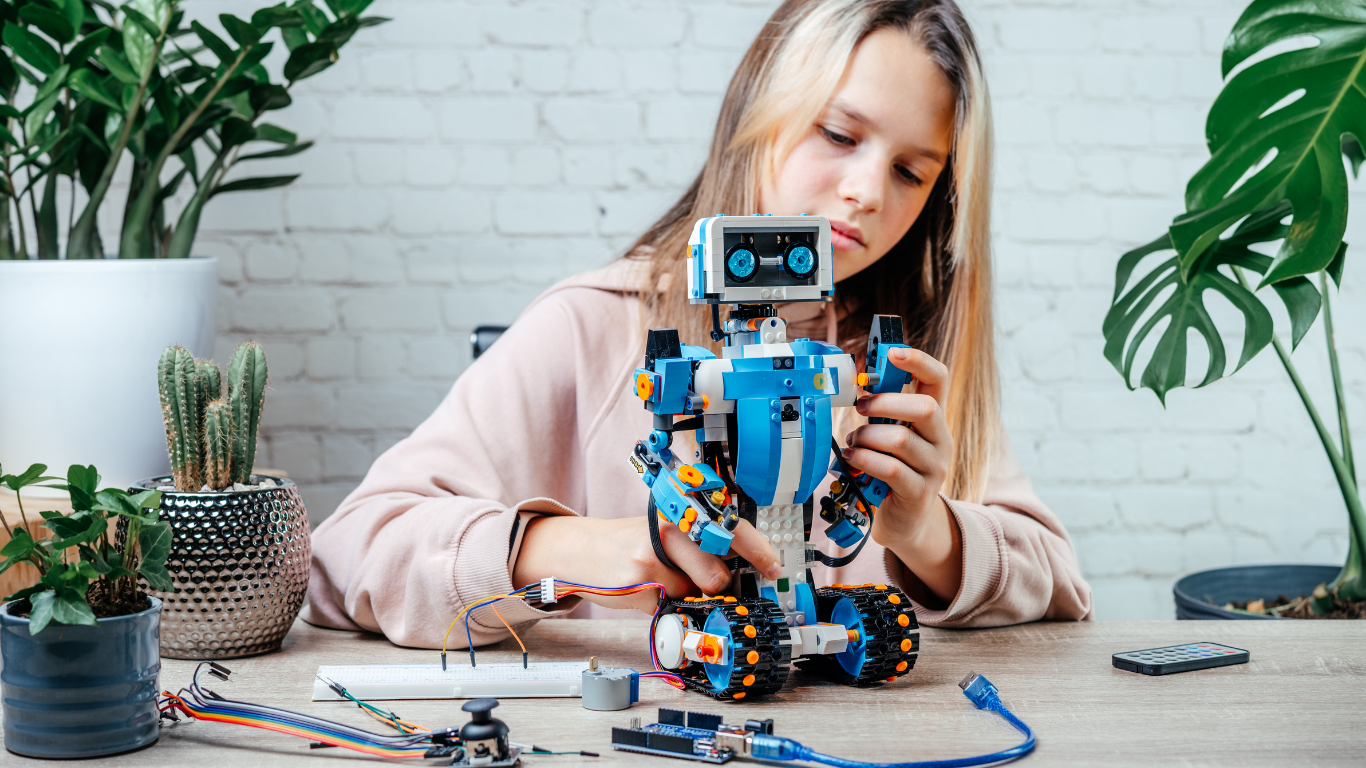 Young girl assembling a blue and white robot with tank-like wheels on a table with electronic components and potted plants.