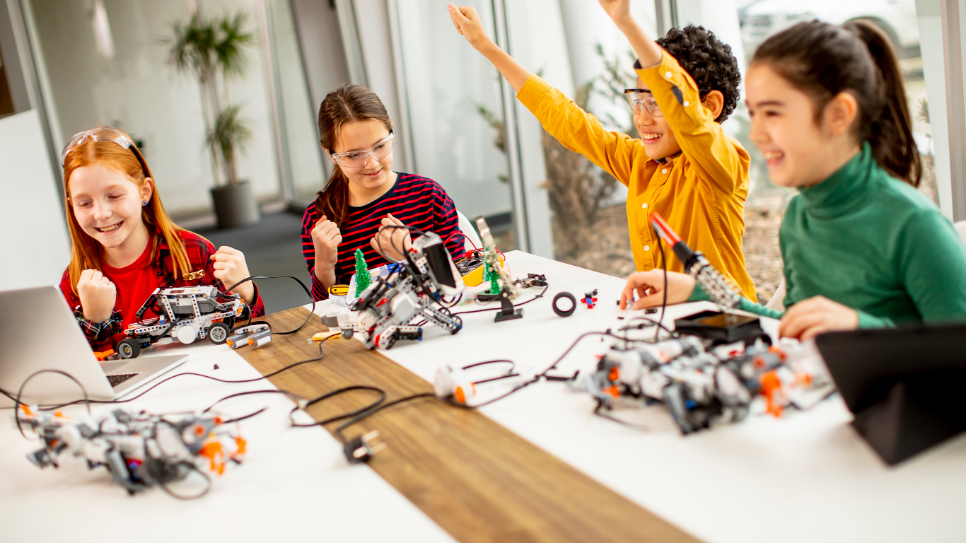 Four children with safety glasses working on and celebrating robotics projects around a table with laptops and robot kits.