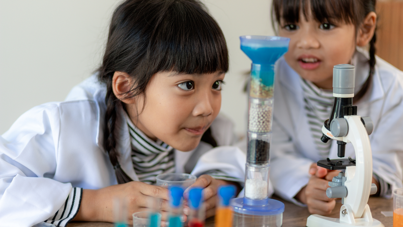 Two young girls in white lab coats observing a layered science experiment and a microscope on a table.