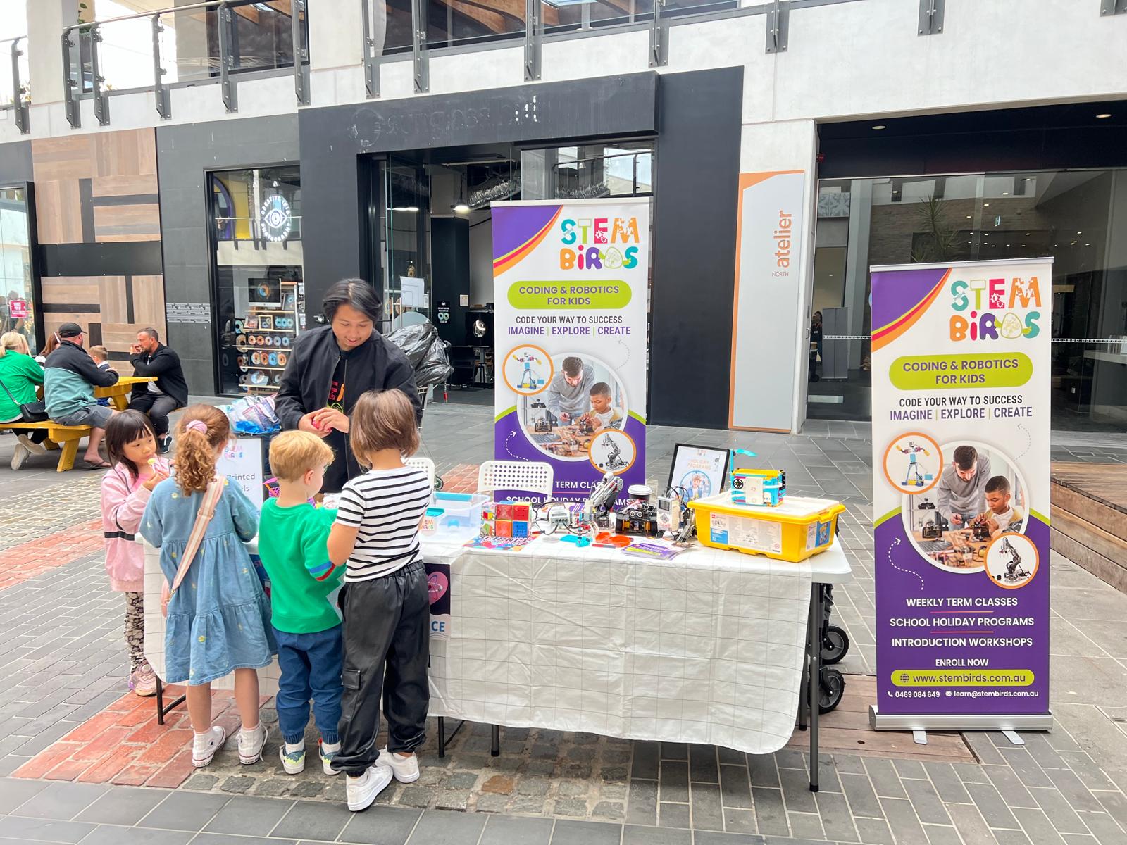 A group of children gathered around a table with robotics kits and a woman explaining STEM Birds coding and robotics programs for kids.