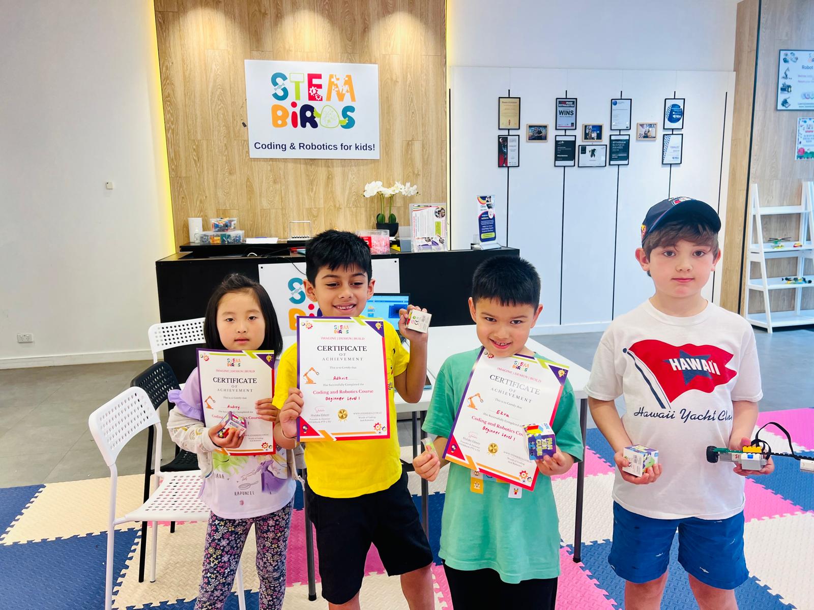 Four children standing indoors holding certificates and robotics kits at a STEM BIRDS Coding & Robotics for kids center.