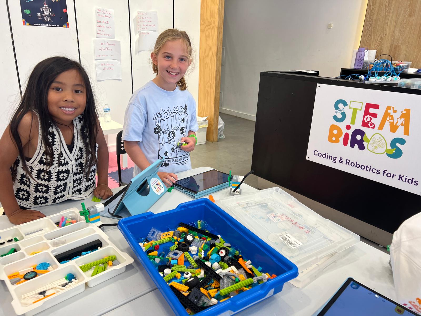 Two young girls smiling at a table with LEGO robotics parts and tablets in a STEM Birds coding and robotics class for kids.
