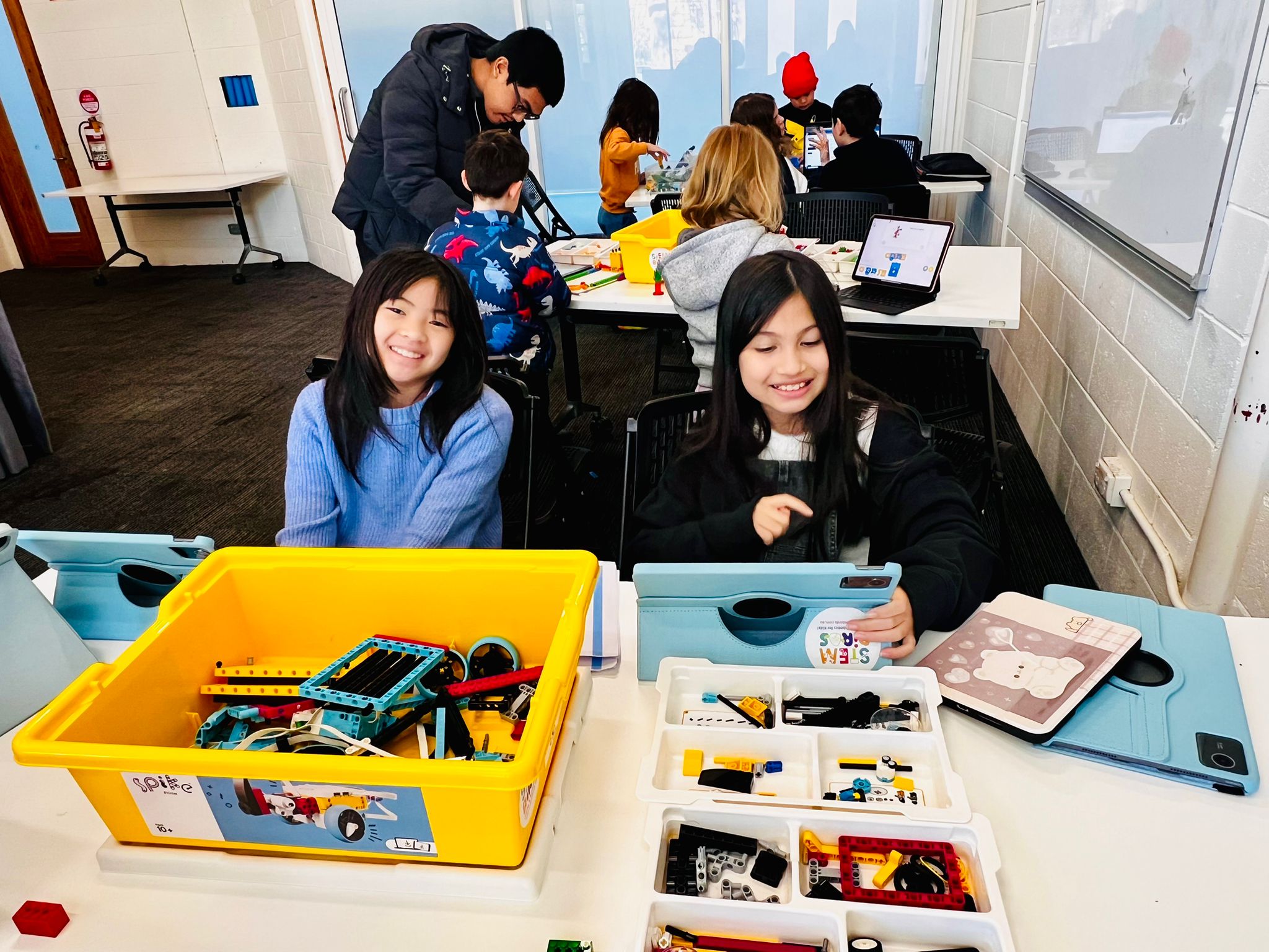 Two girls smiling while sitting at a table with robotics kits and tablets in a classroom with other children working in the background.