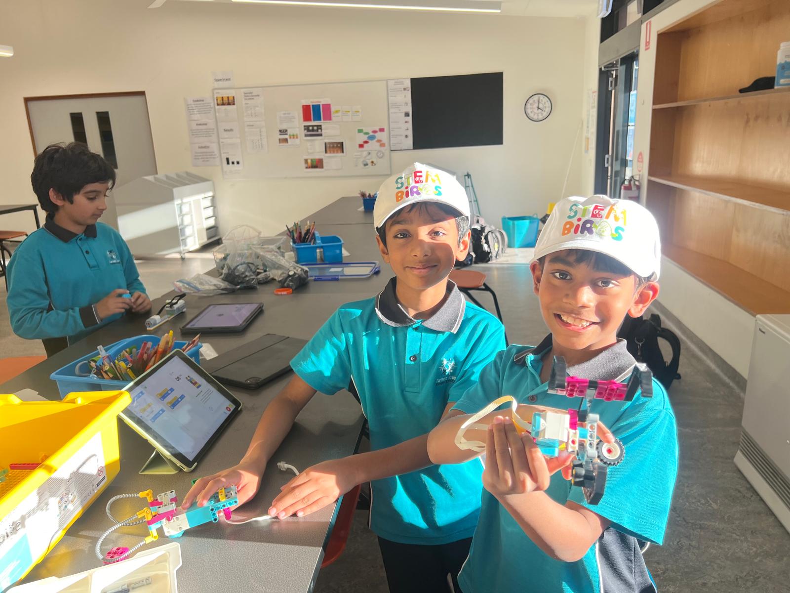 Two boys in teal shirts and STEM Birds caps holding a robotics project at a classroom table with a third boy working in the background.