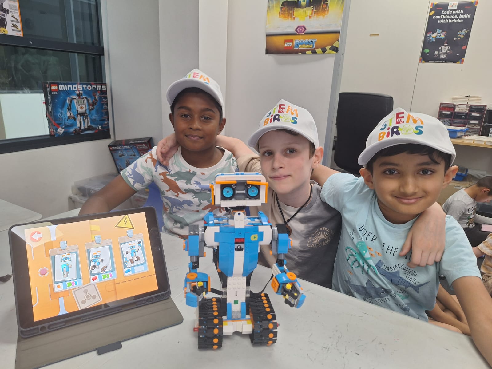Three boys wearing Stem Birds caps posing with a blue and white LEGO robot and a tablet displaying a robot assembly app.