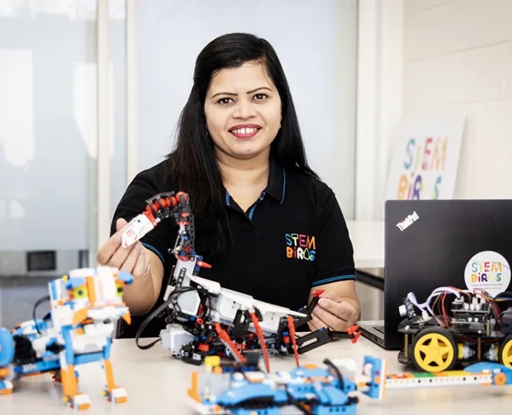 Woman in a STEM Birds shirt smiling while holding a robotic arm surrounded by robotic models on a table with a laptop in the background.
