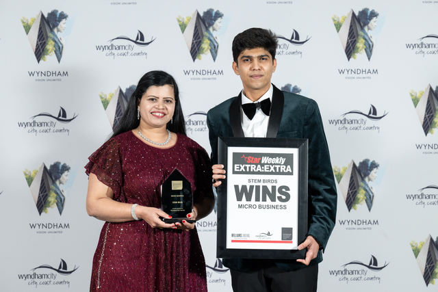 A woman in a sparkling burgundy dress holds a trophy while a man in a dark green velvet suit and bow tie holds a framed award certificate against a Wyndham backdrop.