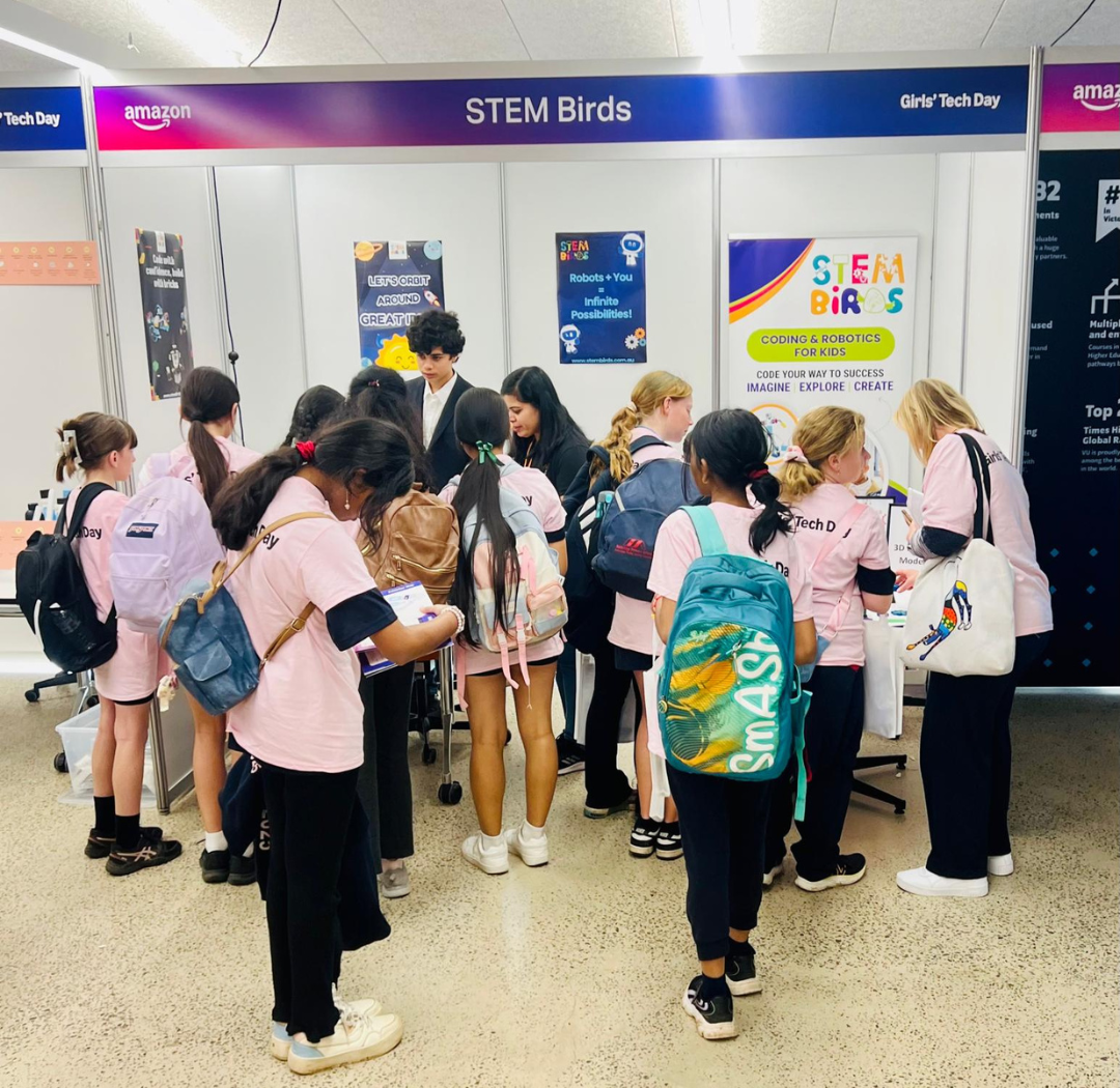 Group of girls wearing pink Girls' Tech Day shirts gathered around a STEM Birds booth with colorful posters about coding and robotics for kids.
