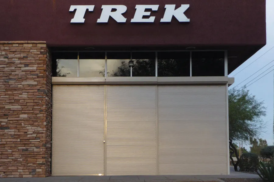 Storefront with "TREK" signage above closed shutters, featuring stone and glass elements, surrounded by greenery.