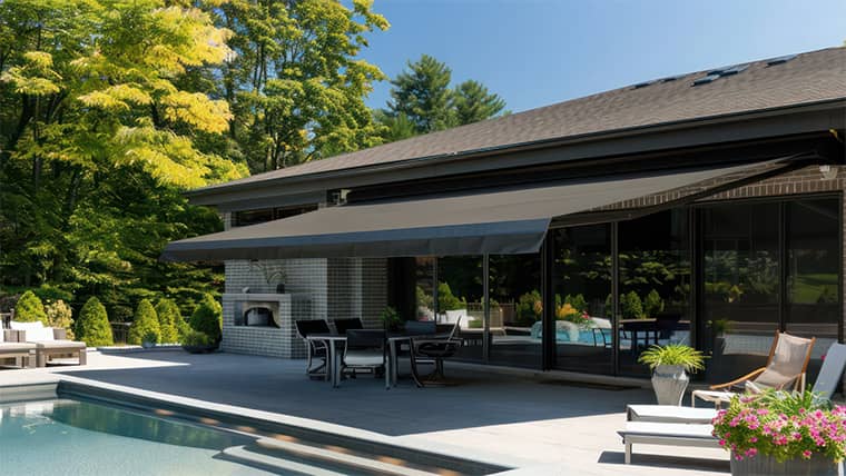 Modern patio with dark retractable awning, outdoor dining set, fireplace, and pool surrounded by lush green trees.