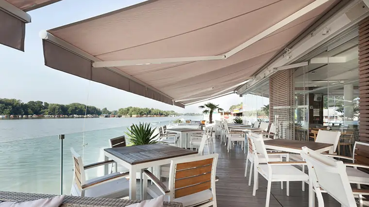 Outdoor dining area with wooden tables and white chairs under large beige awnings overlooking a calm waterfront.