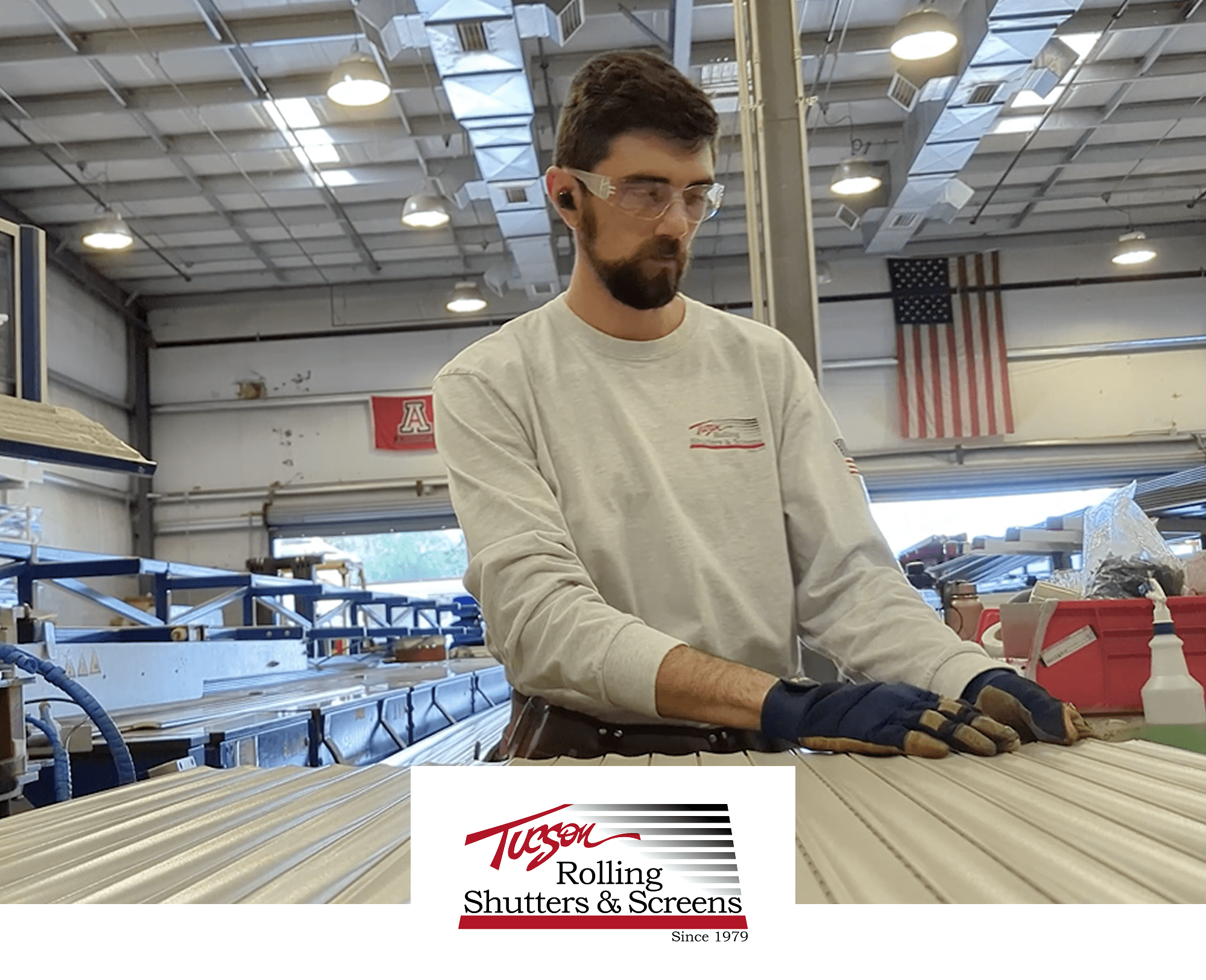 A man wearing safety glasses, gloves, and a Tucson Rolling Shutters & Screens shirt works with metal slats in a factory. The workspace has high ceilings, overhead lights, and American and University of Arizona flags on the wall.