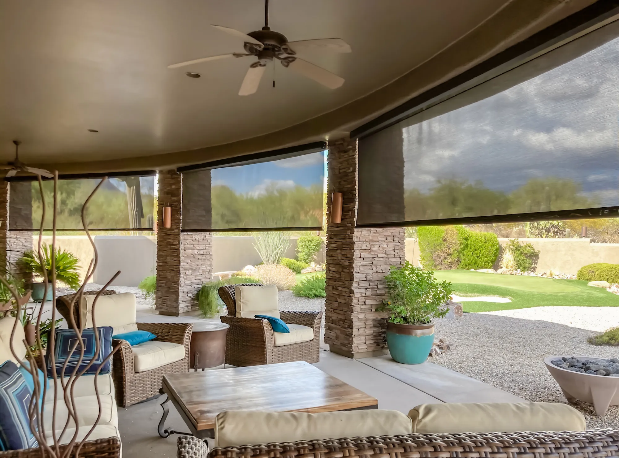 Covered patio with wicker furniture, cushions, ceiling fans, retractable mesh shades, and a view of a landscaped backyard with gravel and greenery.
