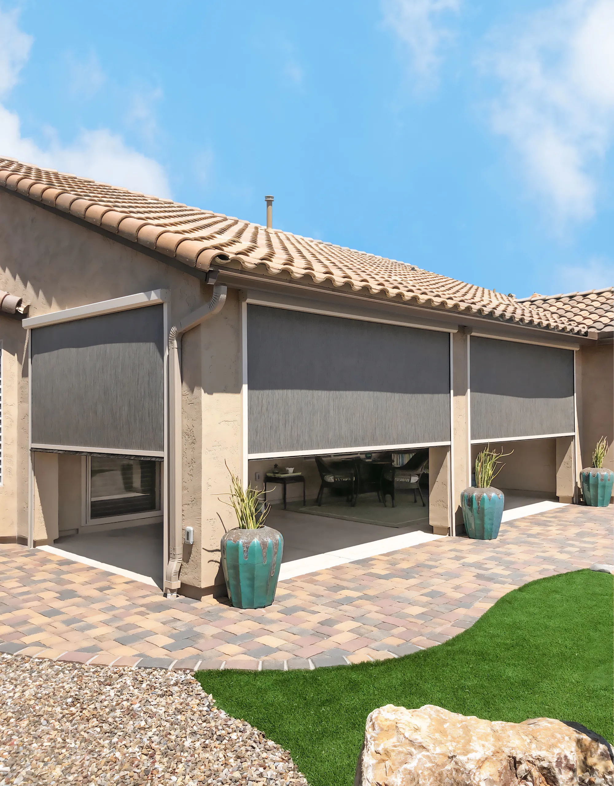 Back patio of a house with three retractable outdoor fabric shades partially lowered, decorative large green pots with plants, stone pavers, and artificial grass under a clear blue sky.
