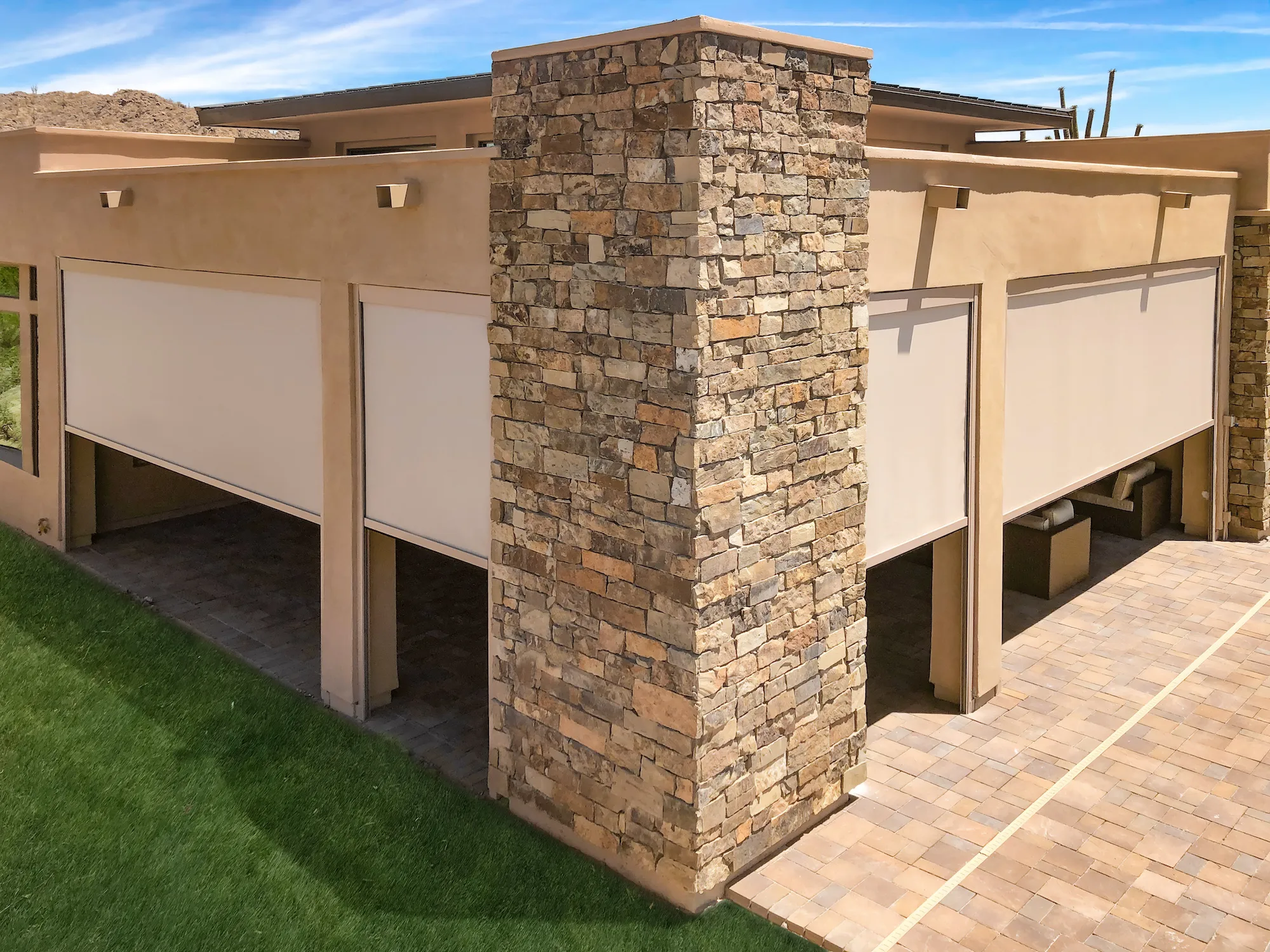 Outdoor patio area with beige fabric roller shades partially lowered, stone column in the center, and seating inside.
