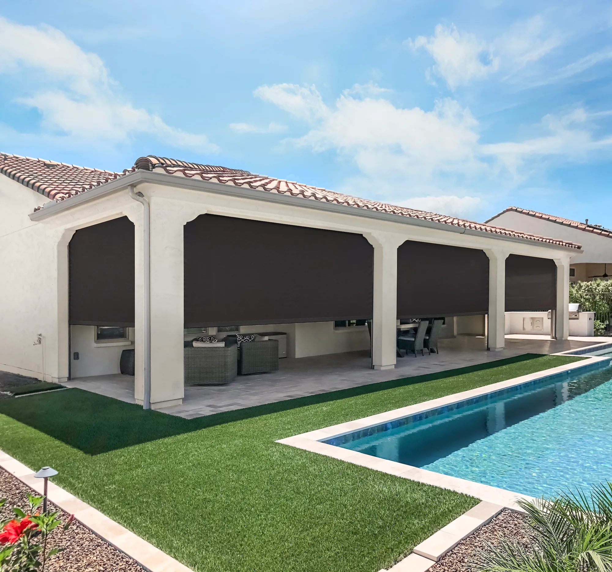Patio area with outdoor furniture behind black fabric roller shades on a house with tiled roof, next to a swimming pool.