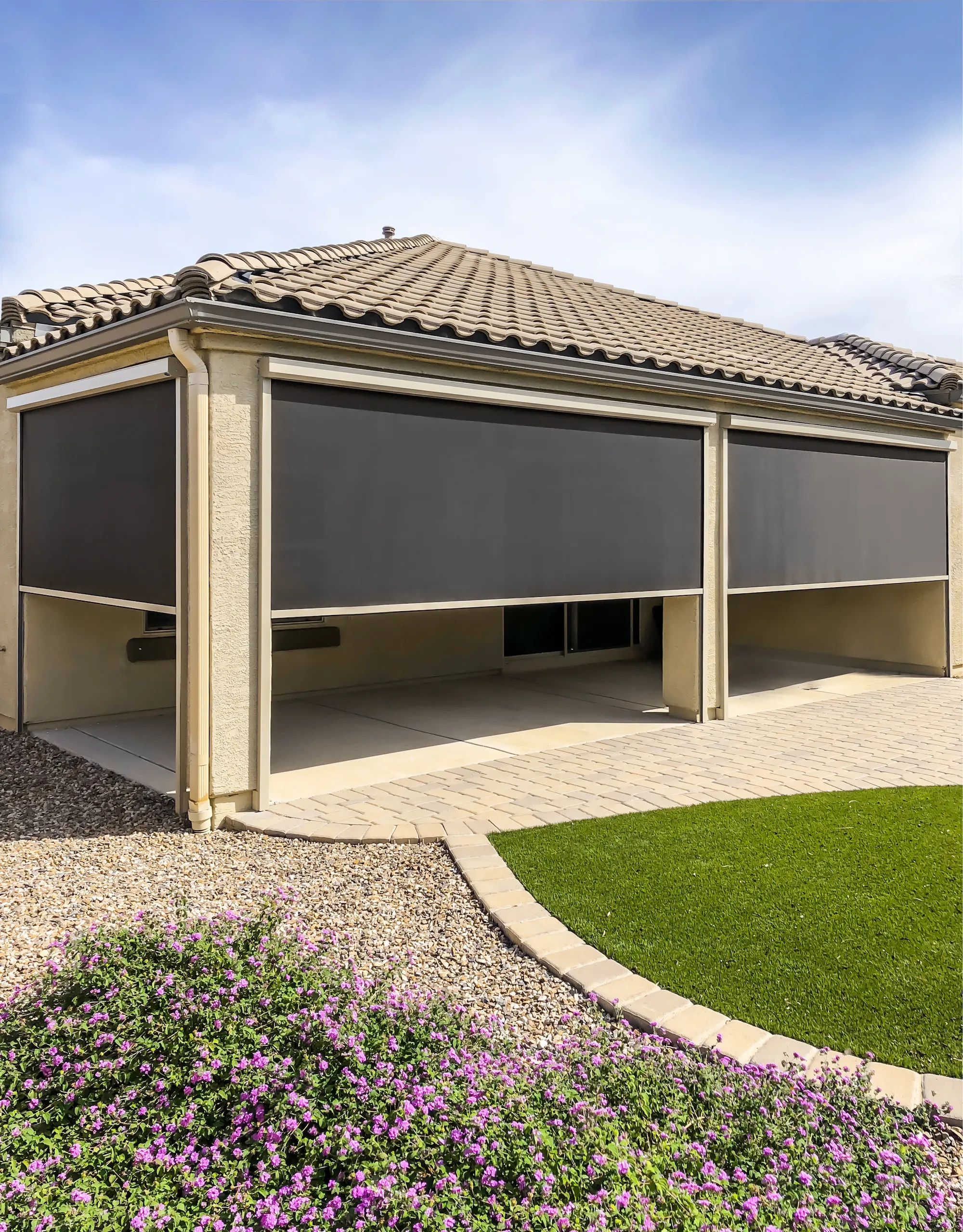 House patio with two large gray outdoor fabric shades partially lowered, surrounded by a stone pathway and landscaped with green grass and purple flowers.