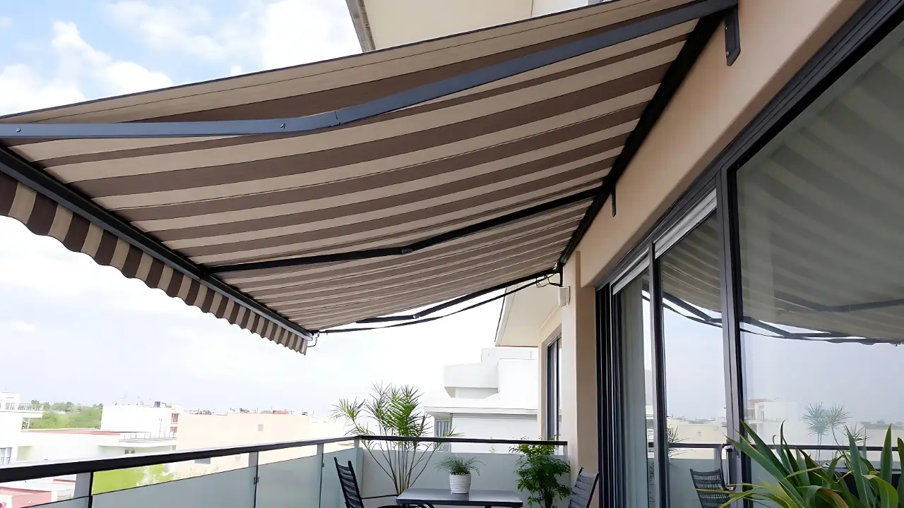 Striped brown and beige retractable awning extending over a balcony with glass railing, outdoor table, chairs, and plants.