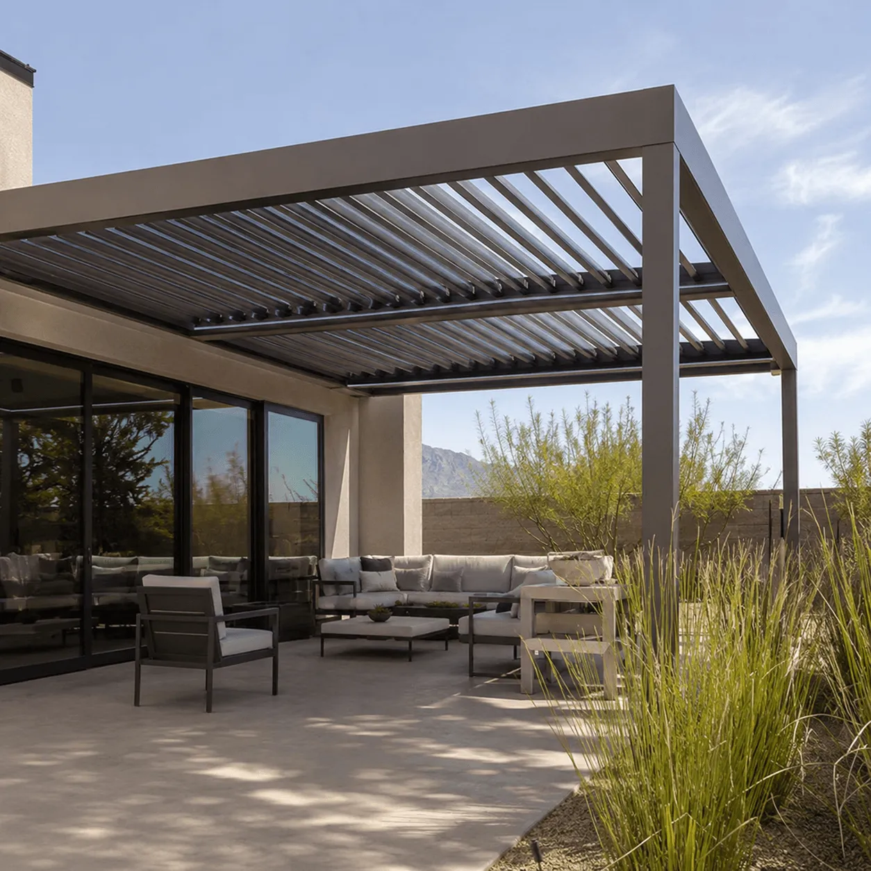 Louvered pergola attached to a house, shading a patio seating area surrounded by desert grasses