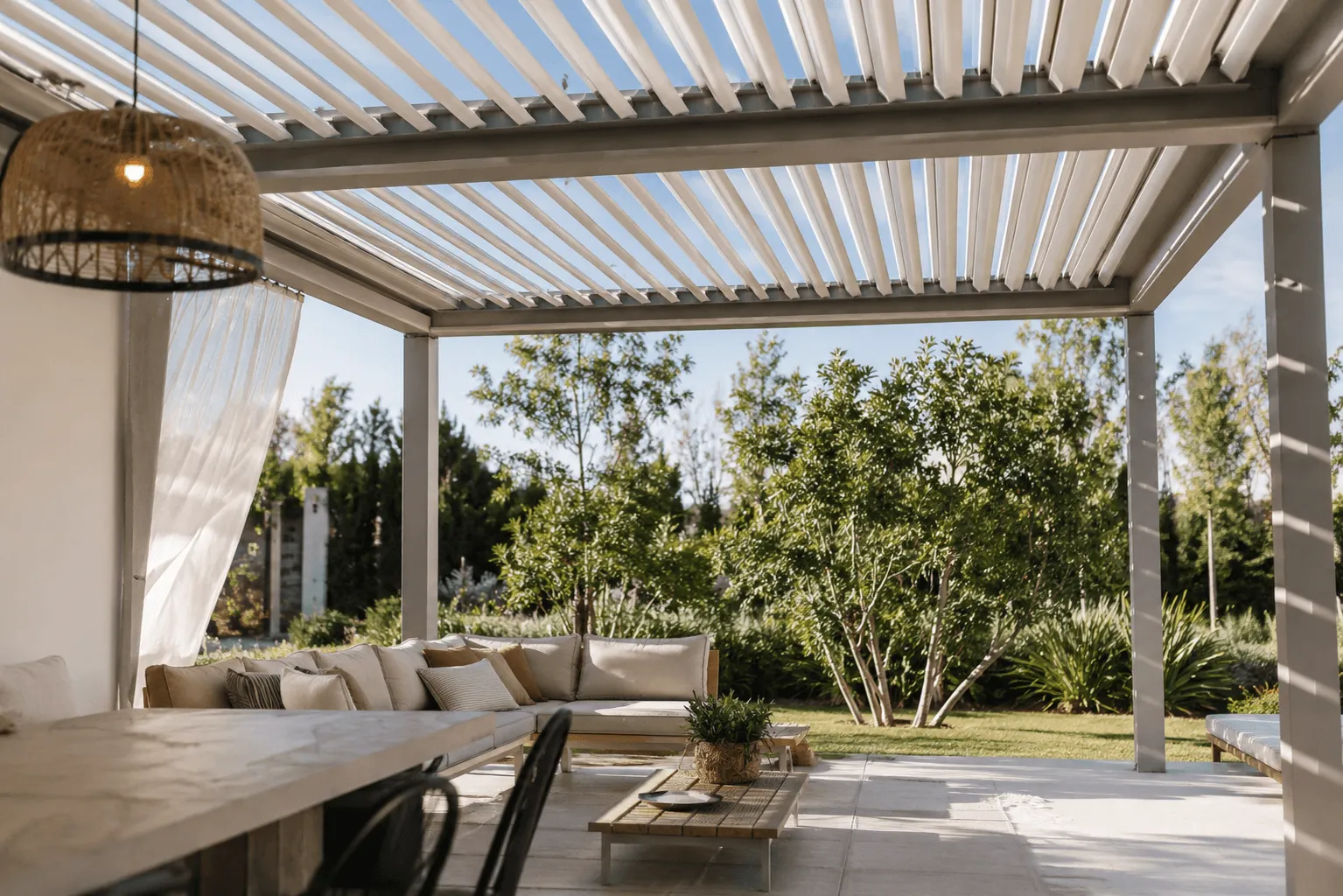 Louvered pergola shading a lounge and dining area with a hanging rattan lamp and sheer curtains, overlooking a garden