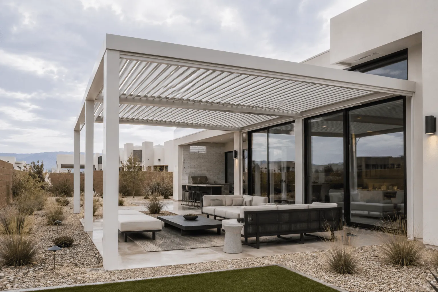 White louvered pergola attached to a modern desert home, covering a patio lounge and outdoor kitchen