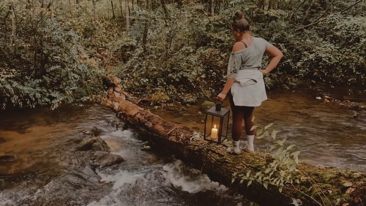 A woman carries a lit candle lantern across a mossy log over a rushing mountain creek