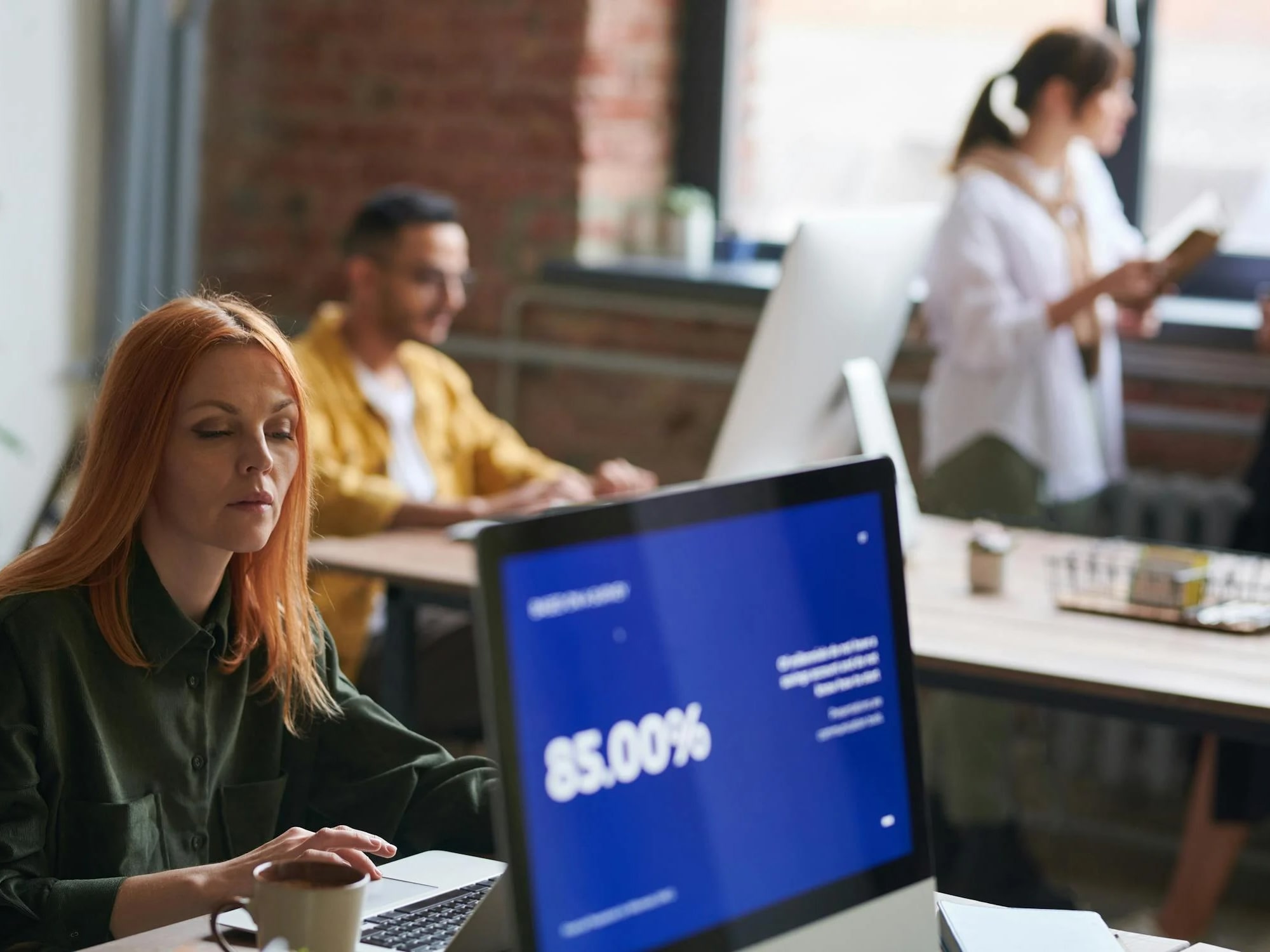 Woman with red hair working on a laptop beside a coffee cup in an office with colleagues in the background.