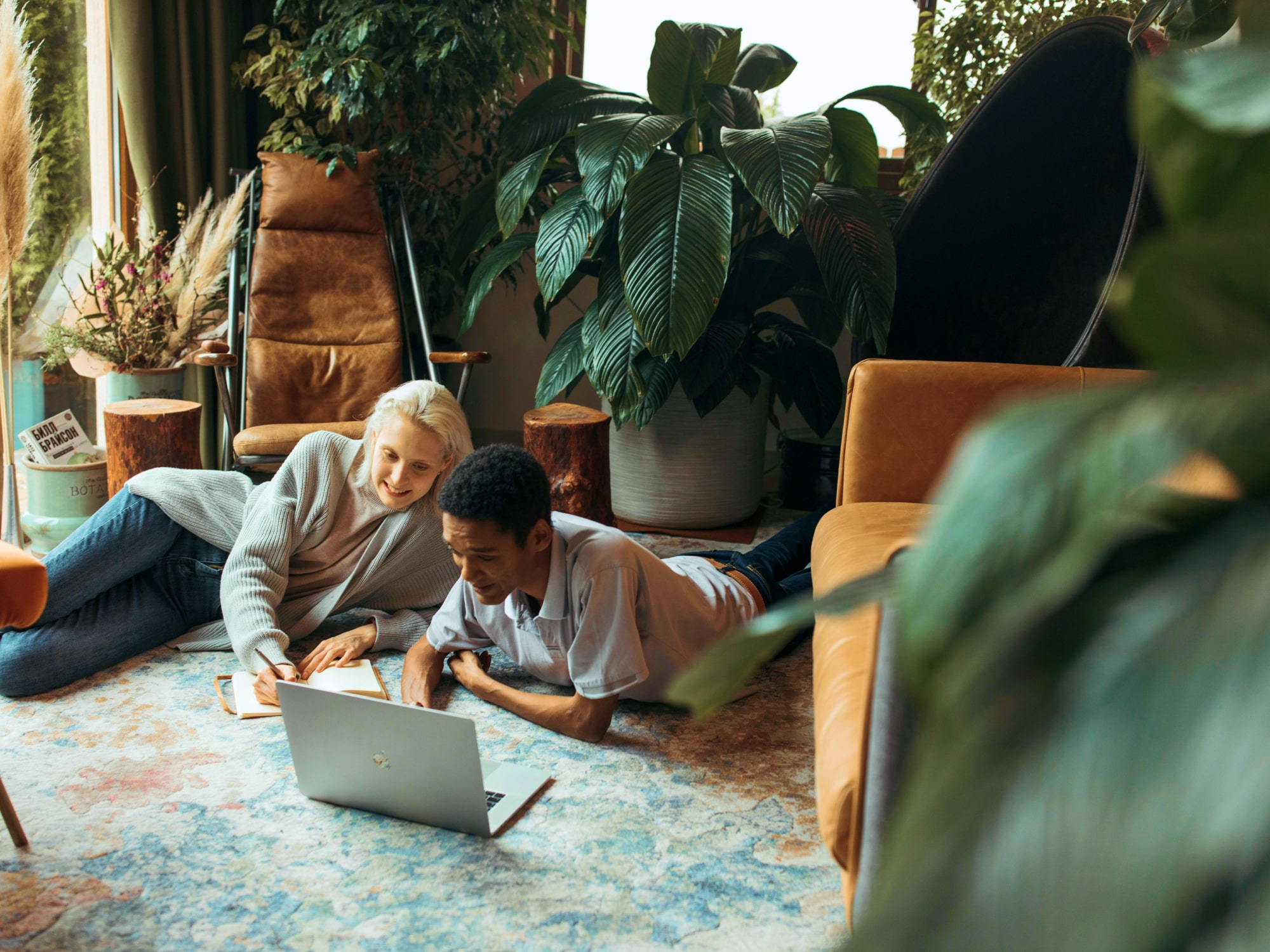 Two people lying on a patterned rug in a cozy room, looking at a laptop and writing in a notebook surrounded by plants and comfortable furniture.