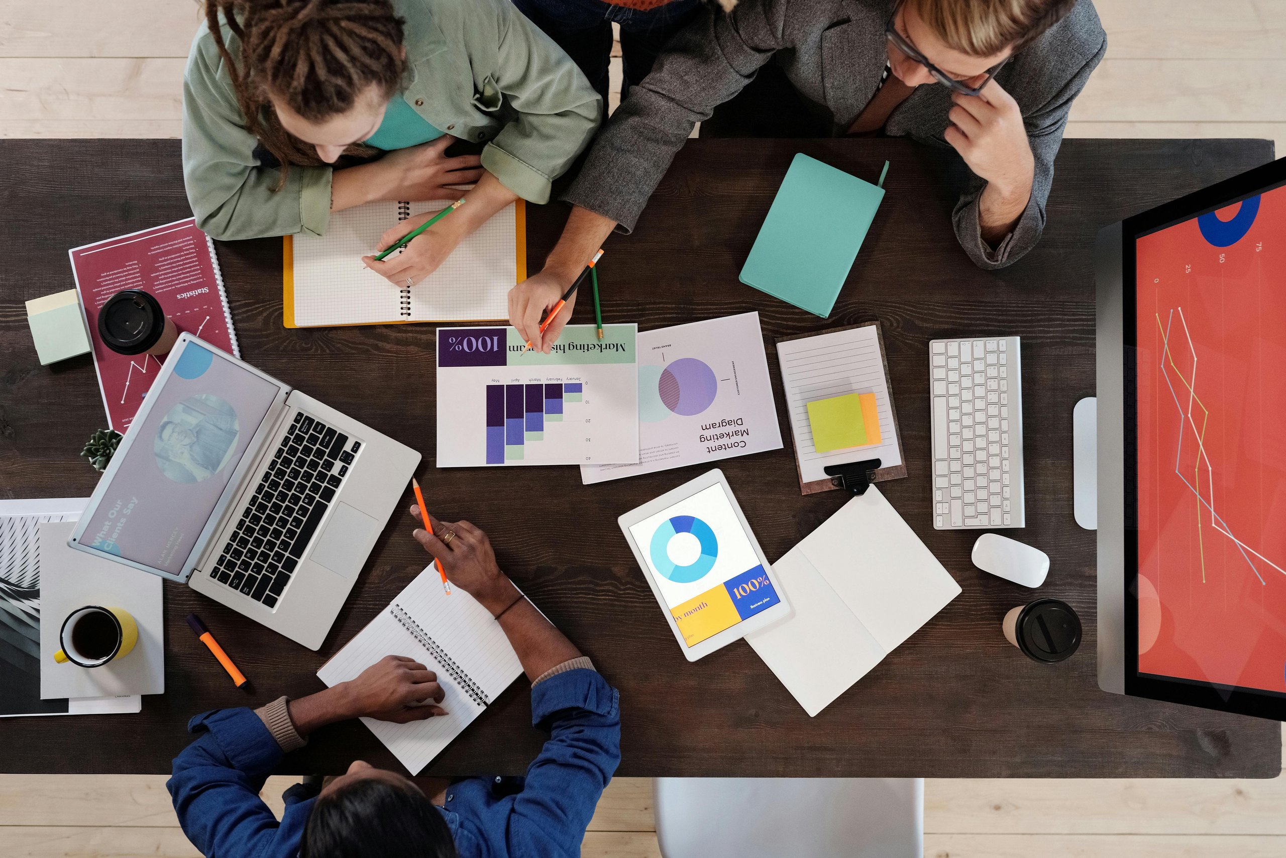Three people collaborating at a table with laptops, tablets displaying graphs, notebooks, and charts.
