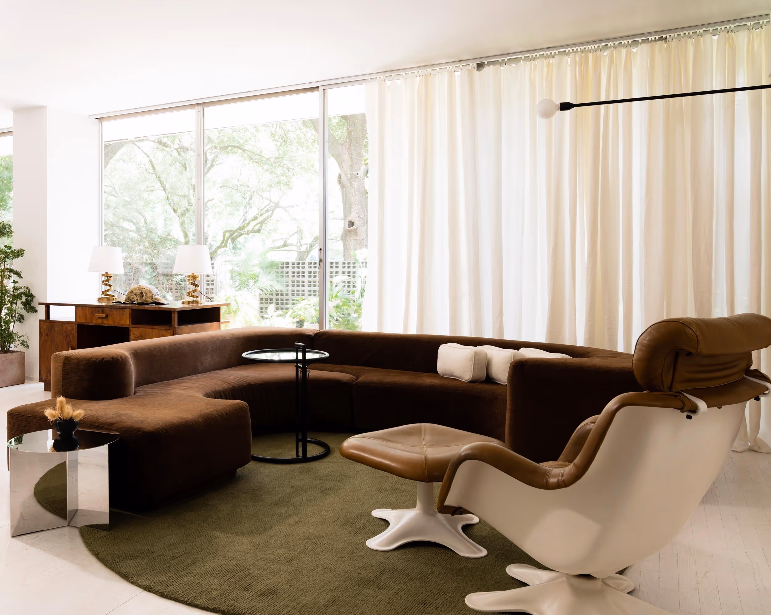 Large dark brown couch on an olive green circle rug, partially open white curtains line the back wall revealing the outdoor space