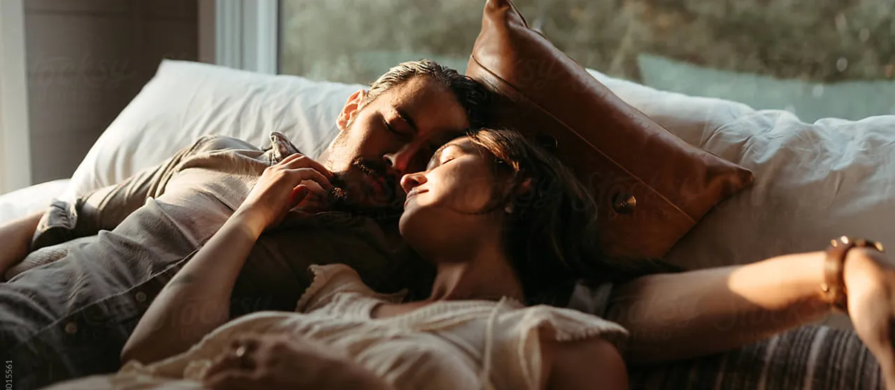 Couple resting closely together on a couch with a brown leather pillow, bathed in warm natural light.