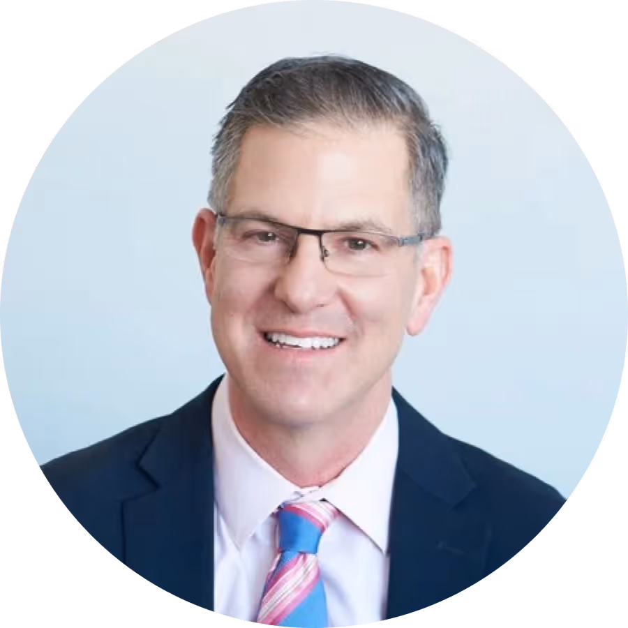 Smiling middle-aged man wearing glasses, a dark suit, light pink shirt, and blue and pink striped tie against a light background.