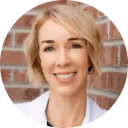 Smiling woman with short blonde hair wearing a white top, standing in front of a brick wall.