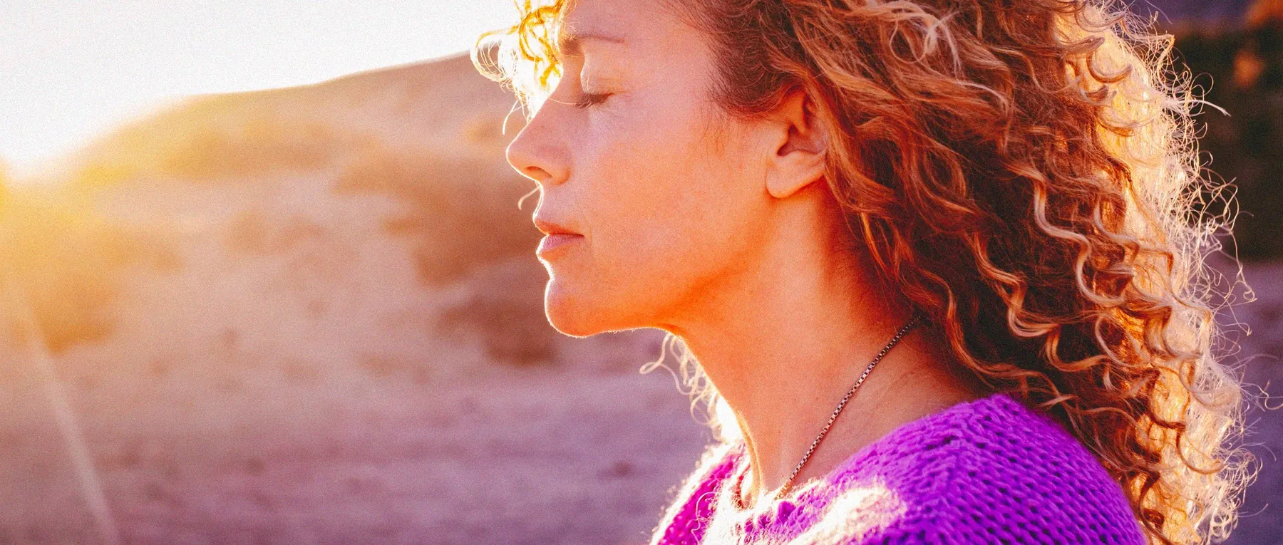 Close-up profile of a woman with curly hair meditating with closed eyes in soft sunset light.