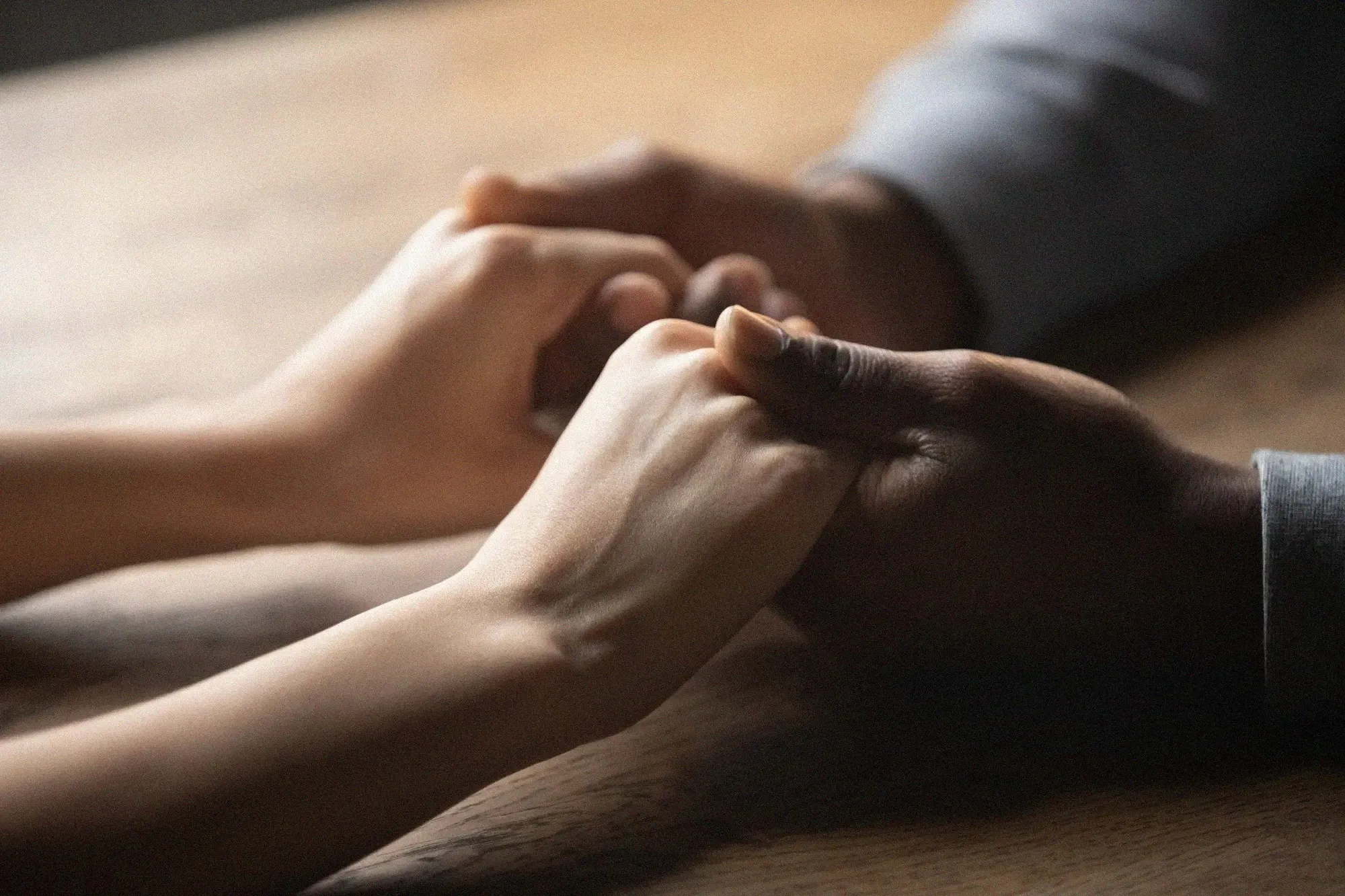 Close-up of two hands, one light-skinned and one dark-skinned, gently holding each other over a wooden surface.