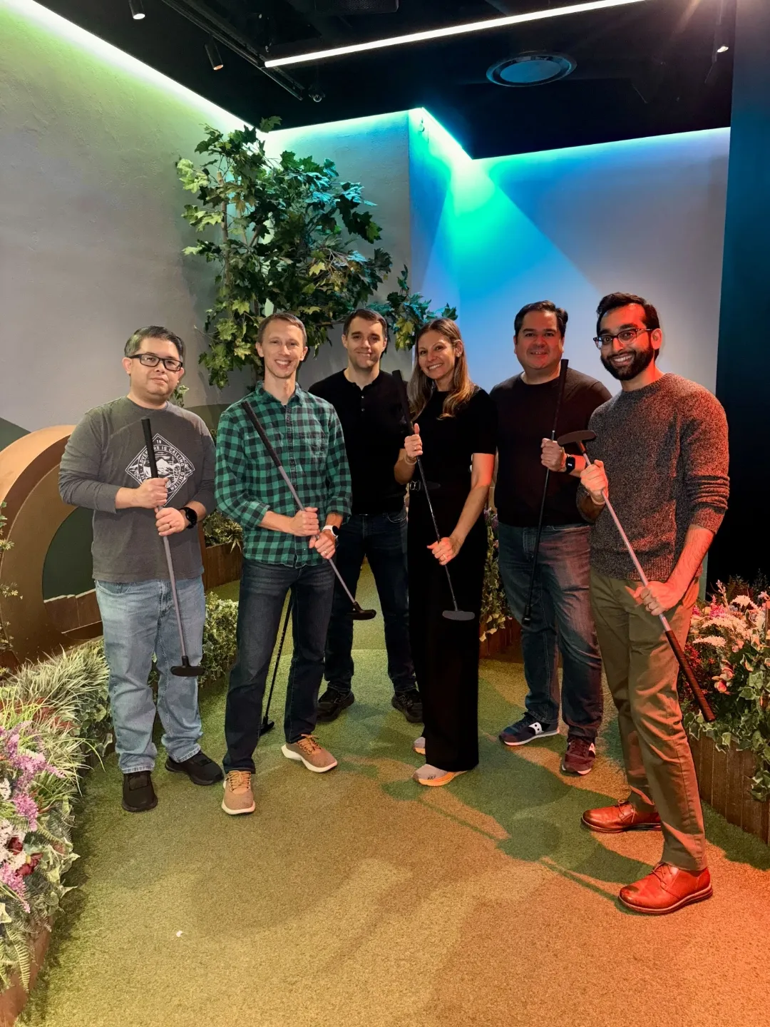 Six adults standing indoors on artificial turf holding mini golf putters, surrounded by plants and colorful lighting.
