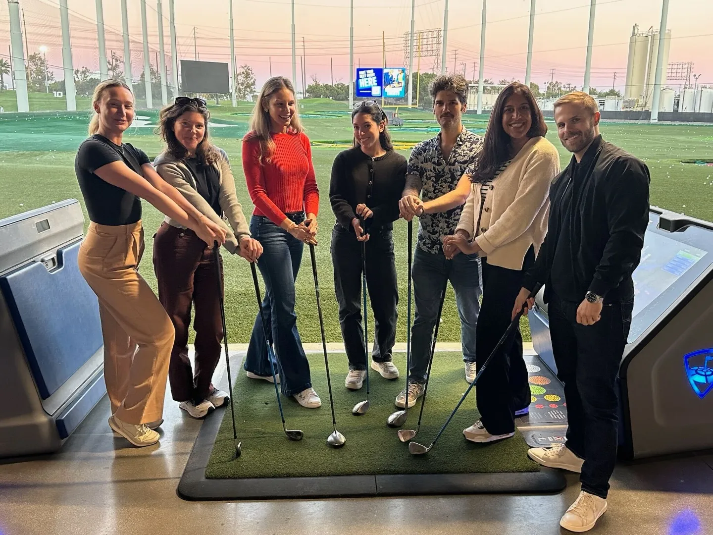 Group of seven people standing on a golf driving range at sunset, each holding a golf club and smiling at the camera.