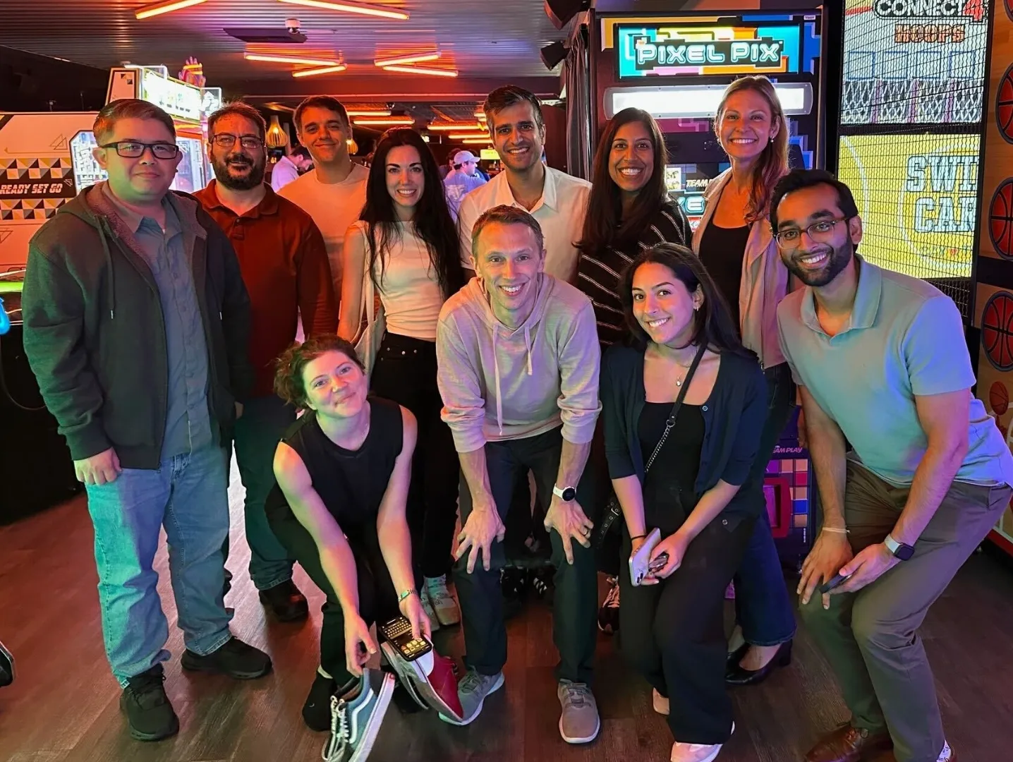 Group of eleven people smiling and posing for a photo in an arcade with games like Pixel Pix and Connect 4 in the background.