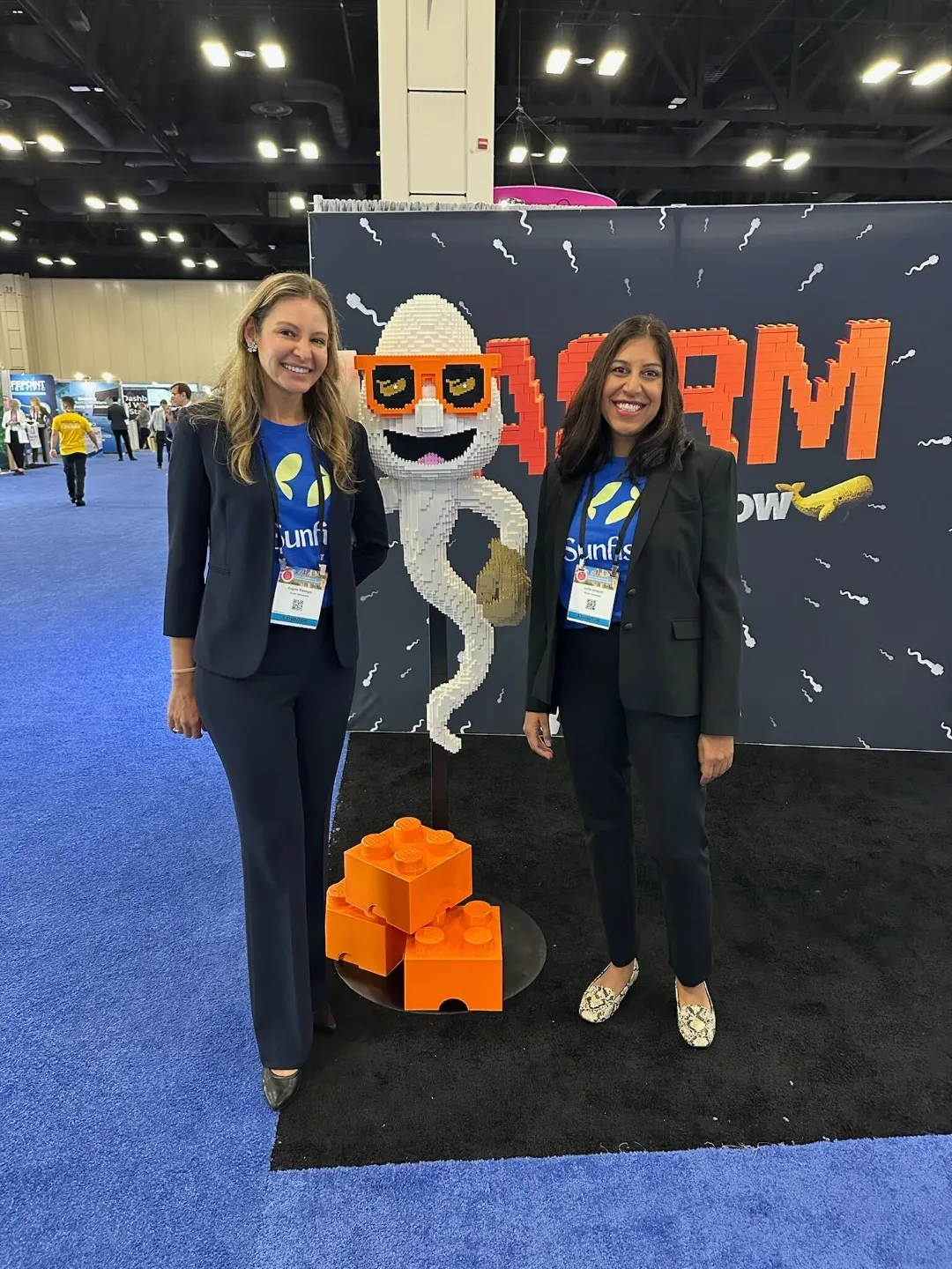 Two women in black suits and blue Sunfish T-shirts stand smiling next to a large LEGO mascot of a sperm with orange sunglasses at a conference booth.