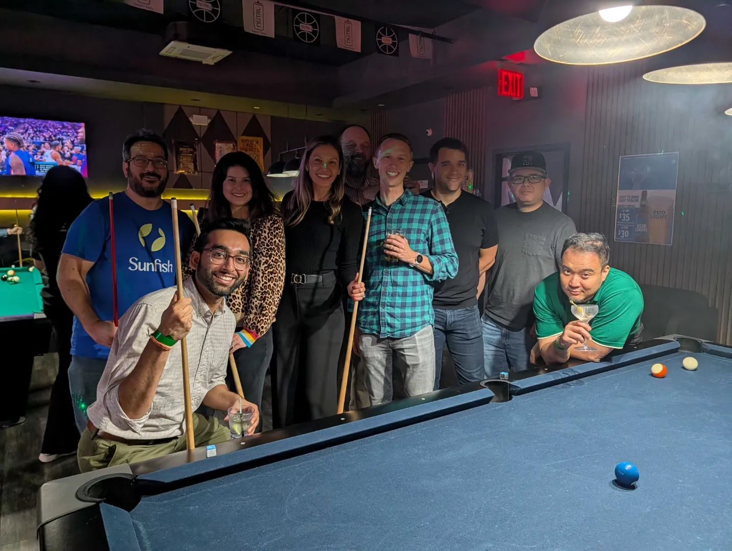 Group of nine people posing with pool cues around a billiards table in a dimly lit room.