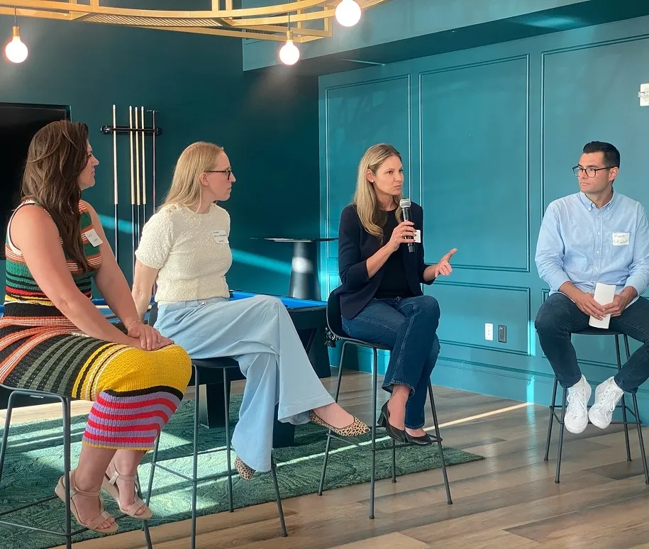 Four adults seated on high stools in a modern room, engaged in a panel discussion with one woman holding a microphone.