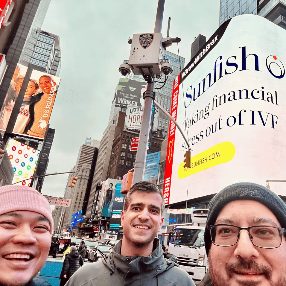 Three men smiling and taking a selfie on a busy city street with tall buildings and a large digital billboard advertising Sunfish and IVF financial stress relief.