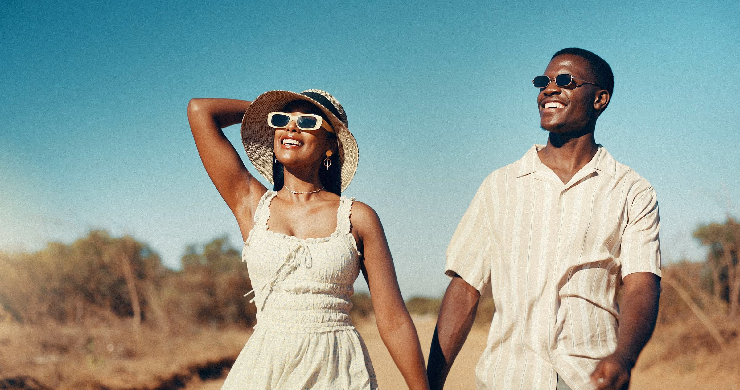 Smiling couple wearing sunglasses, walking outdoors on a sunny day with blue sky background.