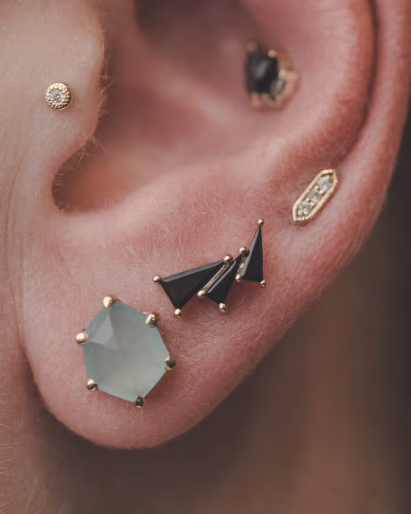 Ear with gold‑framed pale green hexagon gemstone and two black triangular studs plus a small golden accent above – naughtycomrade.ca.